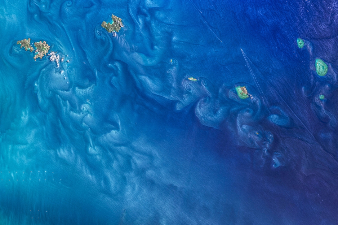 Satellite photo showing the different coloured waters: lighter blue on the left to the darker blues of deeper water on the right, with additional eddy patterns arising in the lee of the islands as the water, and suspended particles, move around them.