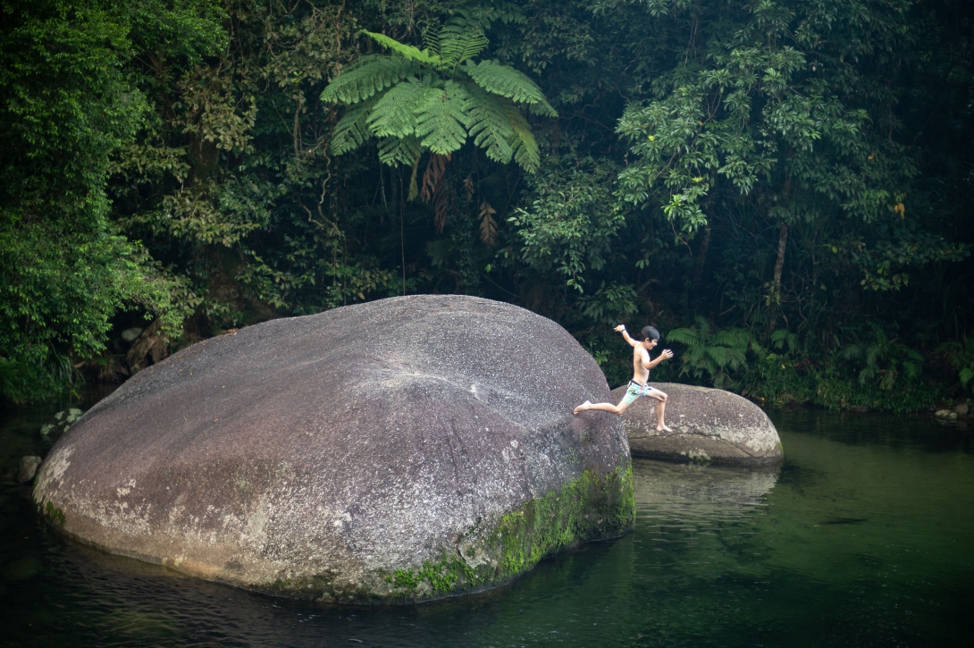 Photograph showing a young boy jumping off a large granite boulder into a clear pool of water that reflects the greens of the rainforest around it. One side of the boulder has moss growing up to about 50cm above the water mark, and lichens cover the other sides near the water. 