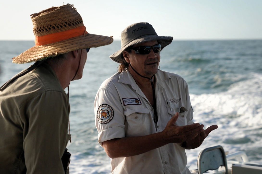 Photograph of two Indigenous rangers in uniform, talking to each other on a boat on the water. The ranger talking has a visible Manduburra ranger badge. 