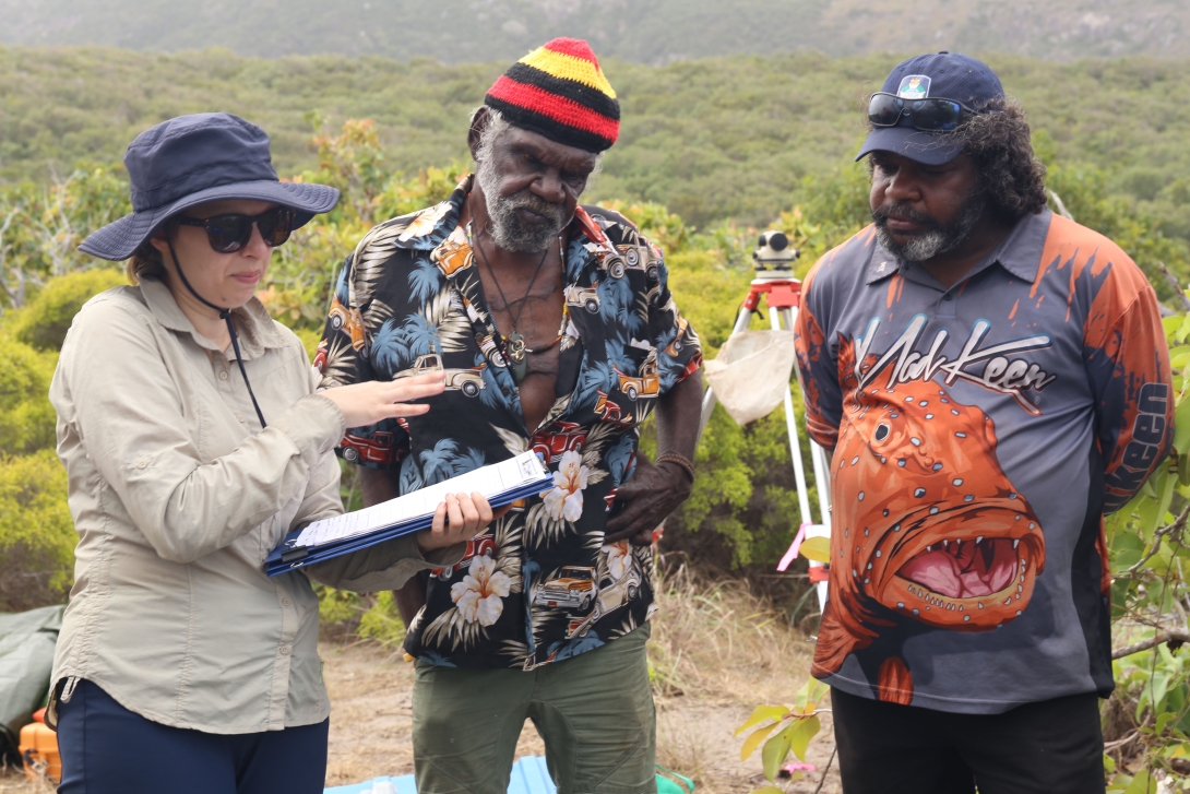 Photograph of a scientist holding several sheets, stacked on top of thin folders, in her left hand while gesturing with her right hand. Two traditional owners stand beside the scientist, listening.