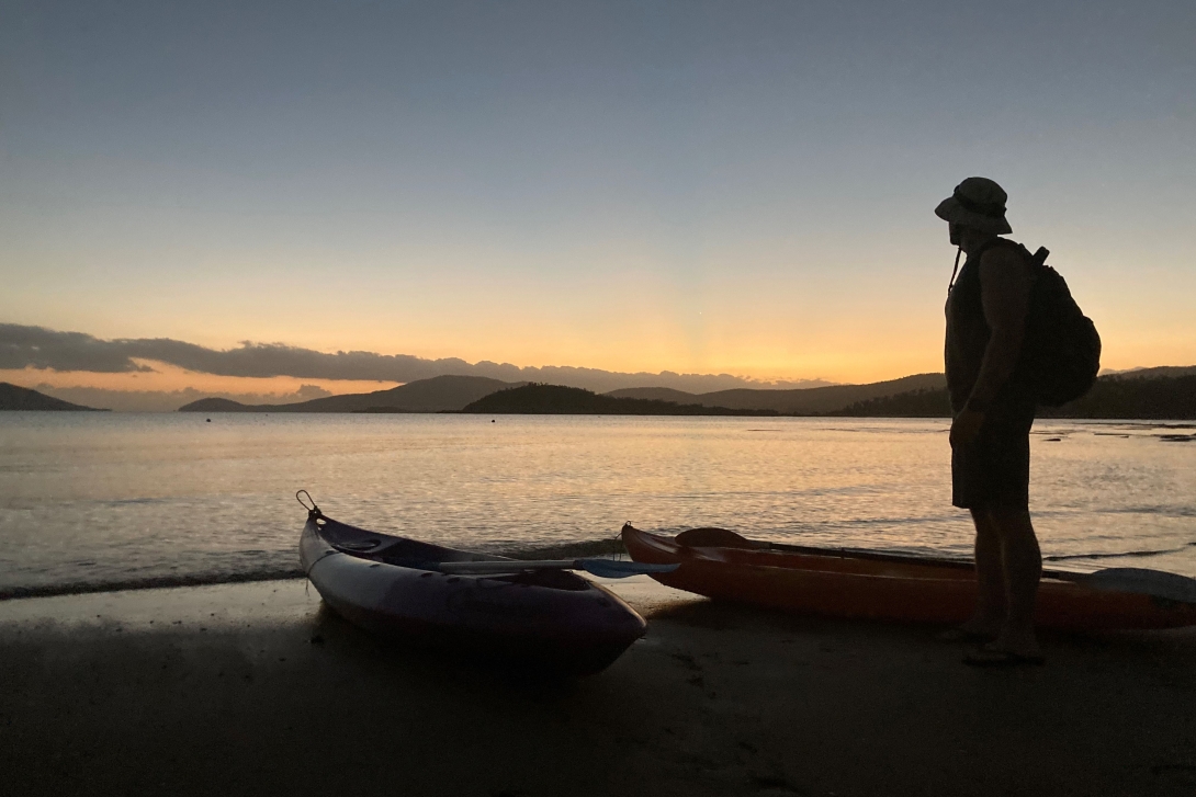 A photograph of the silhouette of a man standing on a beach near the edge of the water at sunrise, looking toward the sea and a mountain range. Two empty kayaks rest nearby.