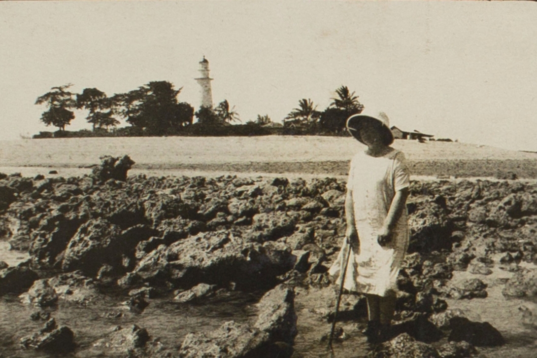 A sepia coloured photograph of a woman dressed in a white mid-length dress and wide brimmed hat, holding a cane. The woman is standing on what appears to be the island’s exposed fringing reef flat. A large part of the island, including its beach, vegetation, lighthouse, and a building, can be seen in the background.