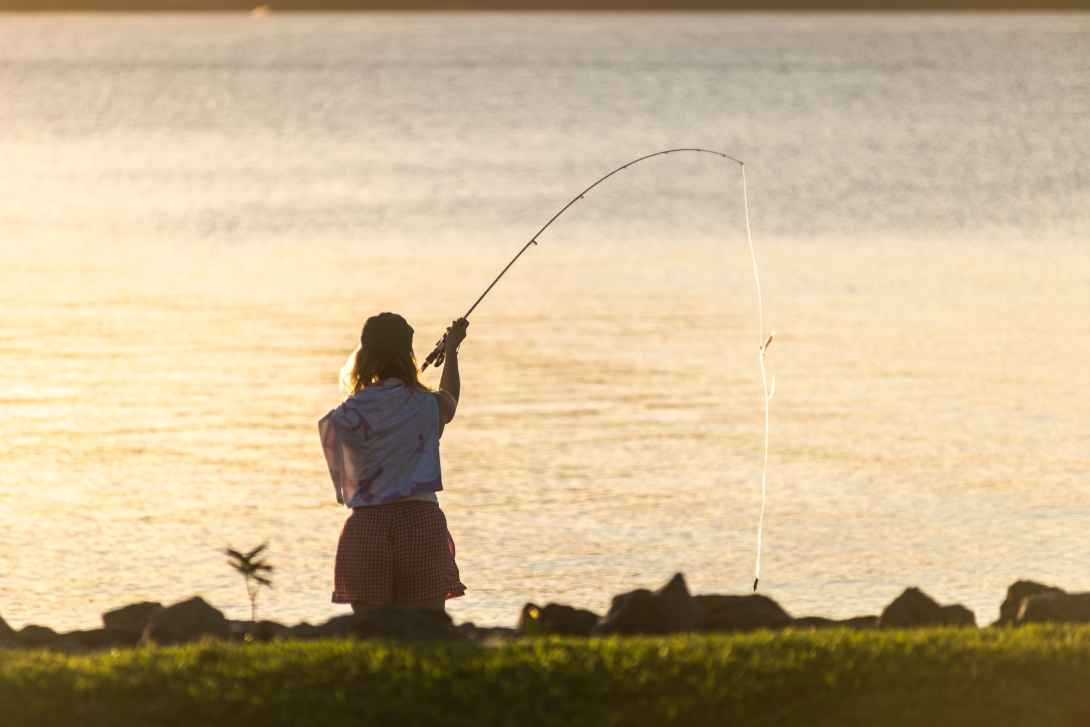 Photo of the back of a woman fishing from a rock wall at sunset. 