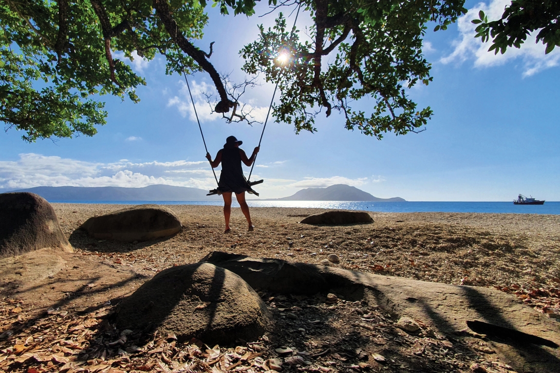 Woman on a swing at the beach looking out towards the ocean, with the sun shining down on the sand and mountains in the far distance. 