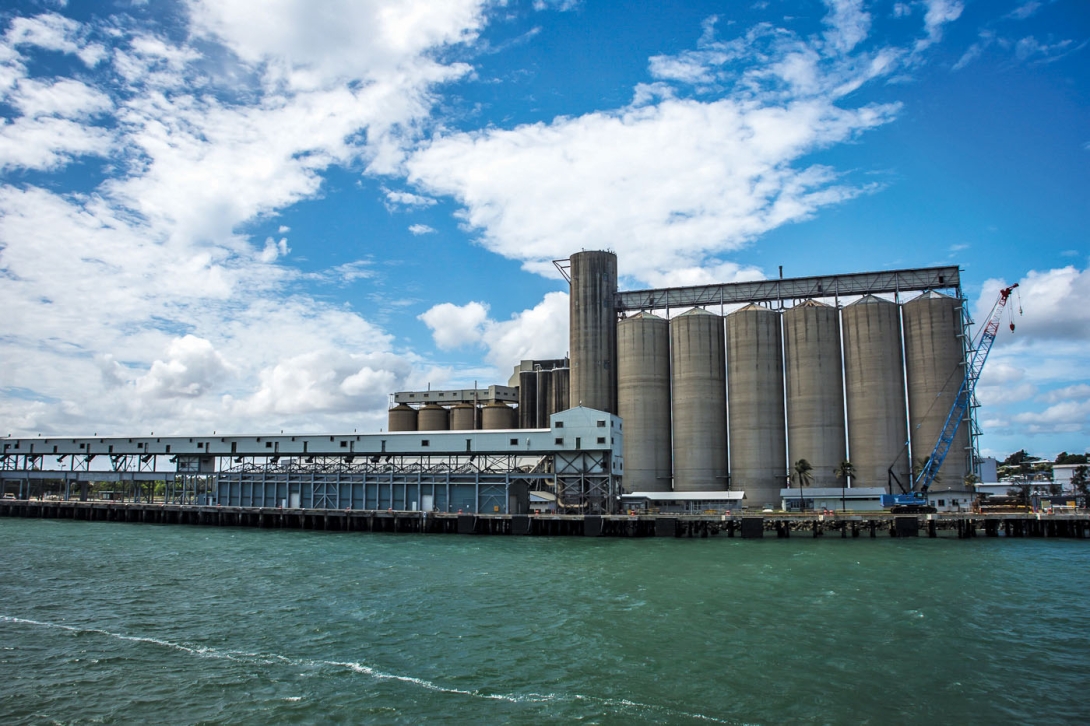 Large concrete storage silos at a Port on the edge of the wharf, with water in the foreground. 