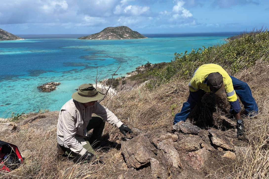 Two men inspecting a pile of rocks with islands and the reef behind them.