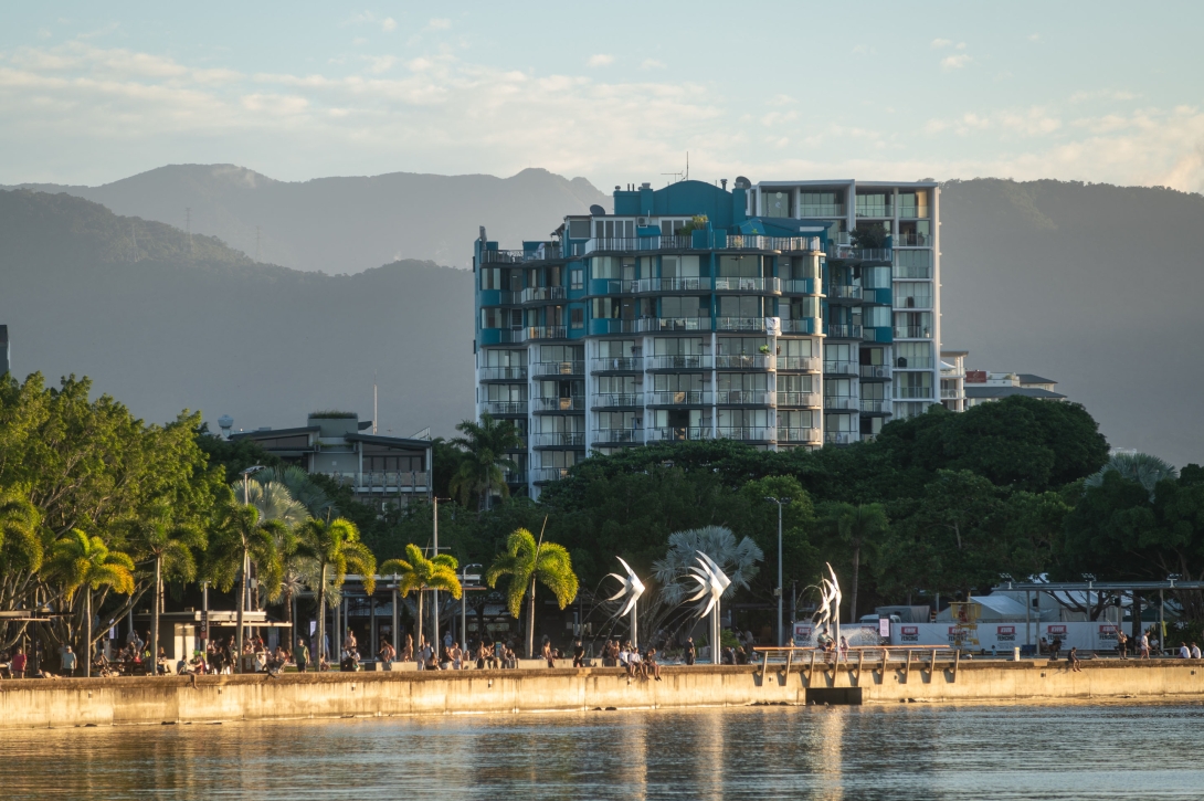 City waterfront with people walking, a high rise building and background of forested hills. The sun shines down on the palm trees and metal statues on the esplanade. 