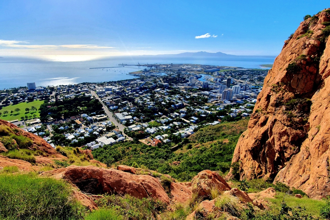 Looking down on Townsville city centre and sea from near the top of Castle Hill, with sunlit red cliff in foreground.