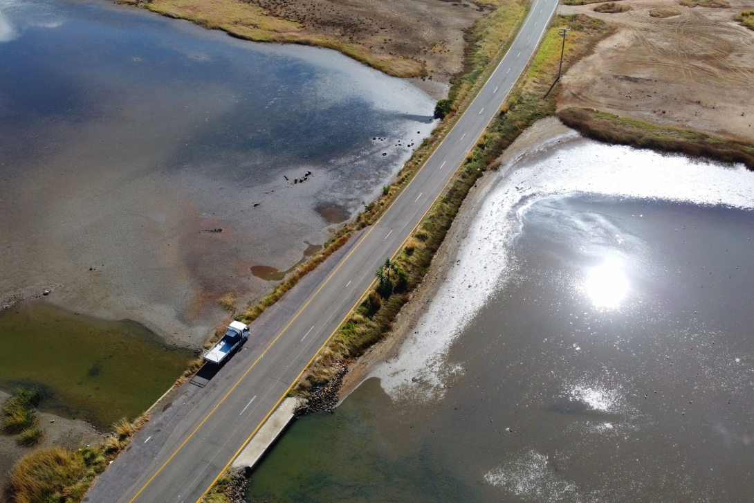 Aerial image of a bitumen road crossing an intertidal wetland, a vehicle can be seen parked on the side of the road. There is an artificial channel letting some water pass underneath the road. 