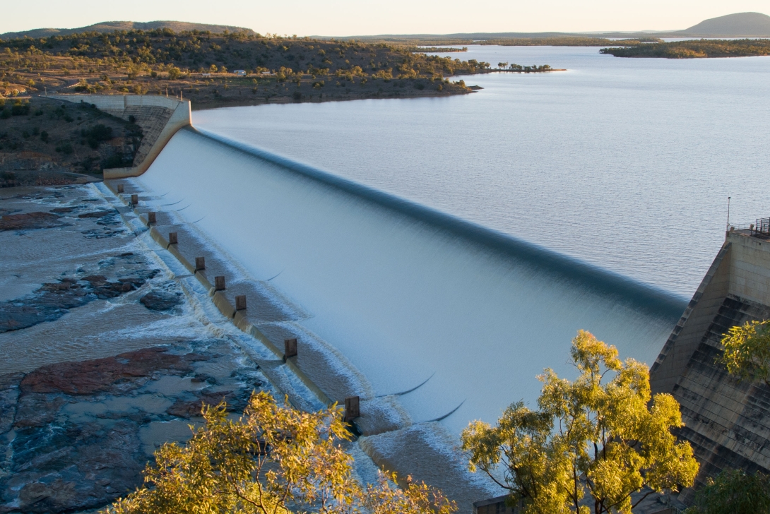 Water flowing over a large dam, with dammed lake and forested landscape behind. 