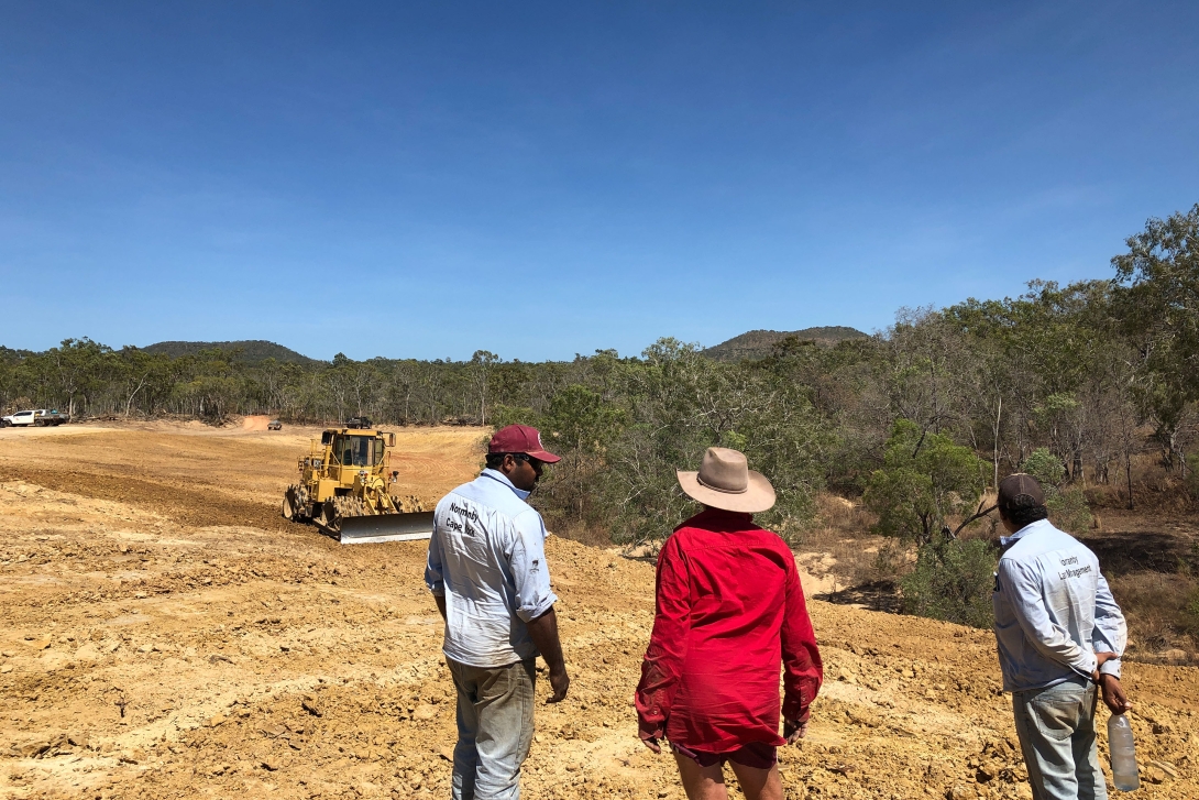 Three people looking across a large area of bare ground where a bulldozer is operating, with a wetland and native forest in the background