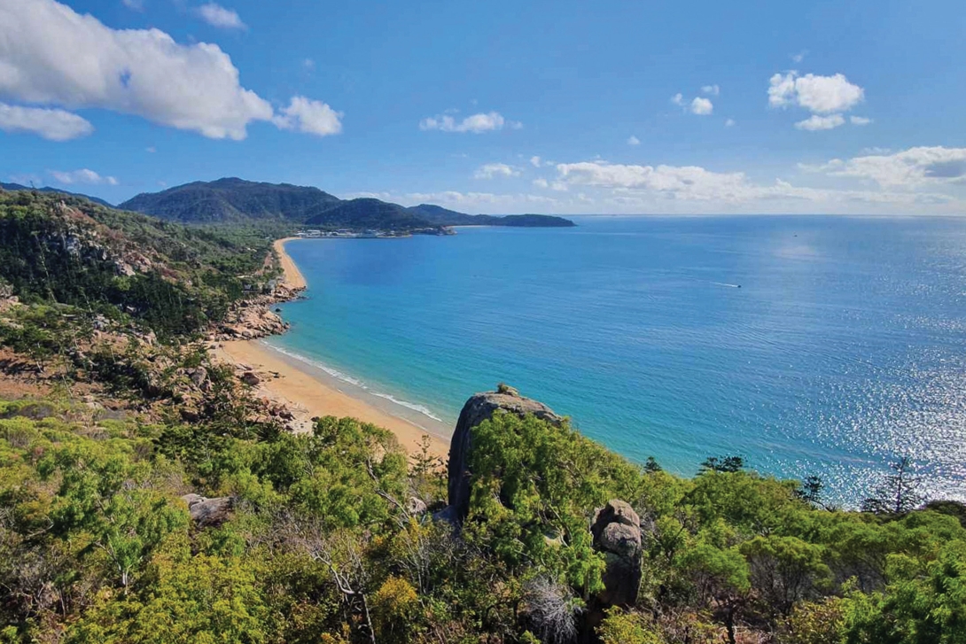 A photo looking out along the length of two beaches on Magnetic Island – Rocky Bay in the foreground and Nelly Bay in the background. There is green coastal vegetaion to the left of the beaches and the blue waters of the Reef to the right. 