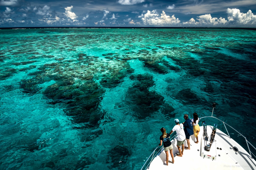 A photo from the top deck of a boat showing the four people standing on the front deck of the boat looking out to an aqua and dark blue coral reef scene, with blue skies and fluffy white clouds in the background over the horizon. 