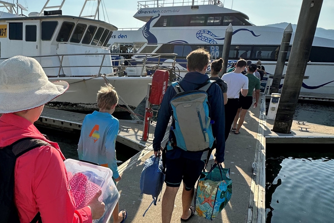 Photograph of a marina with large tourism vessels in the background. Several tourists are waiting in line on the marina wharf to get on a vessel for a visit to the Reef. The tourists are carrying small daypacks.