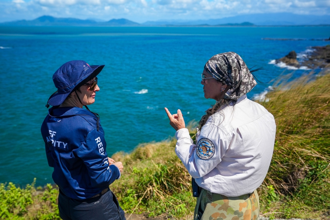 A Reef Authority ranger and a Manduburra Land and Sea ranger discuss something while standing on a grassy cliff edge and looking out to the ocean. 