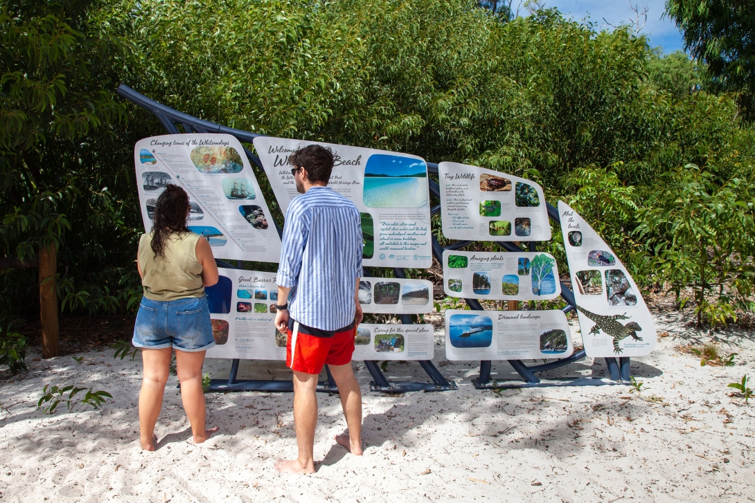 Photograph of two people standing near the beach and reading information about the Whitsundays from a fish-shaped interpretive sign.