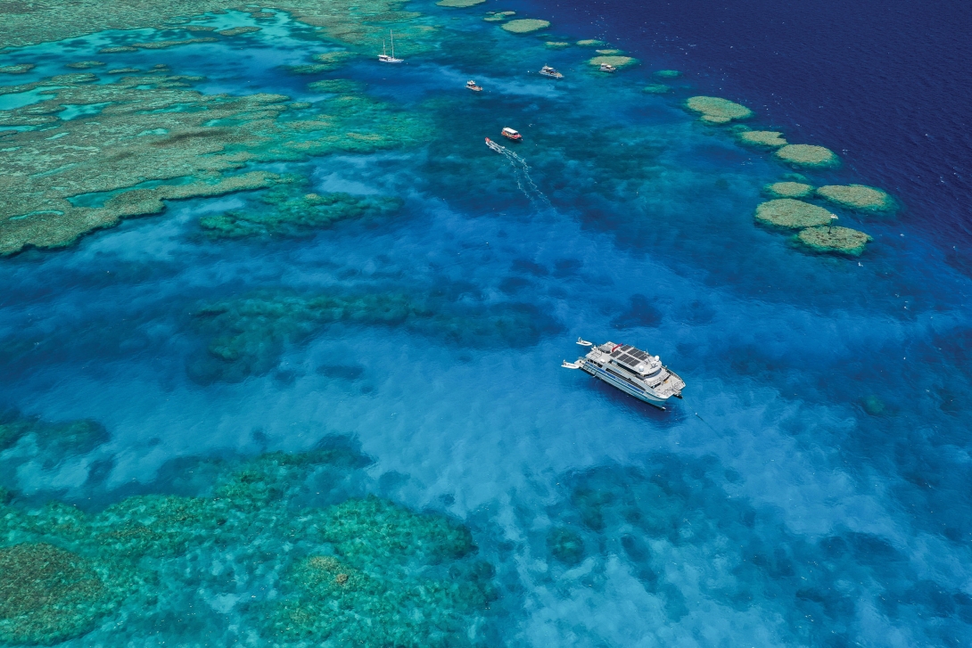 An aerial photograph looking down on coral reef scape – with a large Marine Parks vessel in the foreground, and several other users of the Reef in the top part of the photo. 