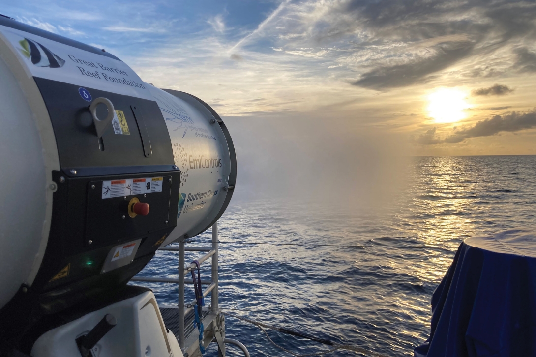 Close-up photograph of a cloud-brightening machine that is directed towards the sea and expulsing a small cloud of water vapour, with a view on the sea, horizon, and sun.