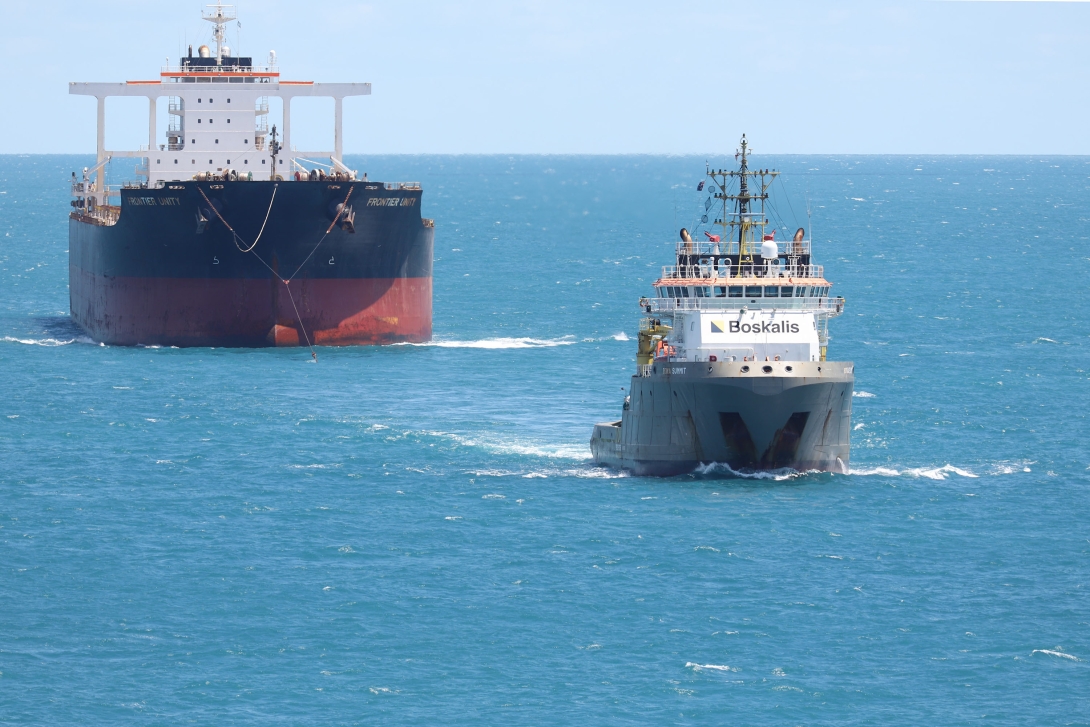 Photograph of a tugboat, pulling a large cargo ship with a tow line towards port.