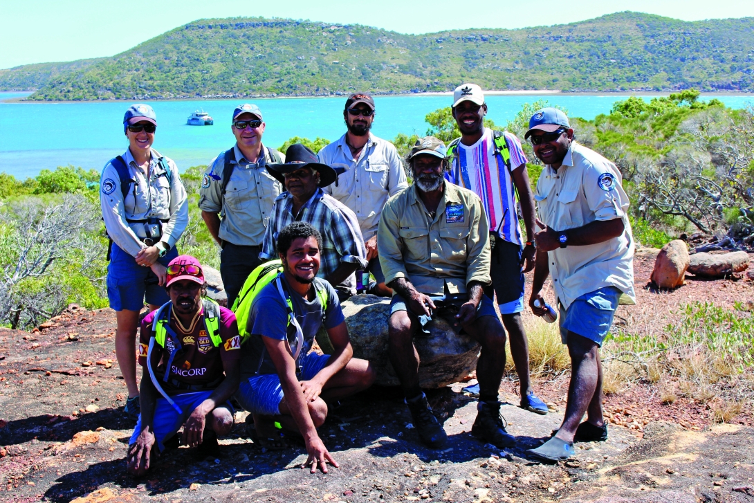 Group photograph of Traditional Owners and Queensland Parks and Wildlife Services rangers. Mountains, the sea, and a vessel can be seen in the background.