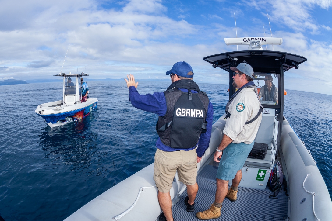 Three rangers from the Reef Joint Field Management Program are standing in a Rigid-hulled inflatable boat, approaching and greeting two people that can be seen onboard a small fishing vessel.