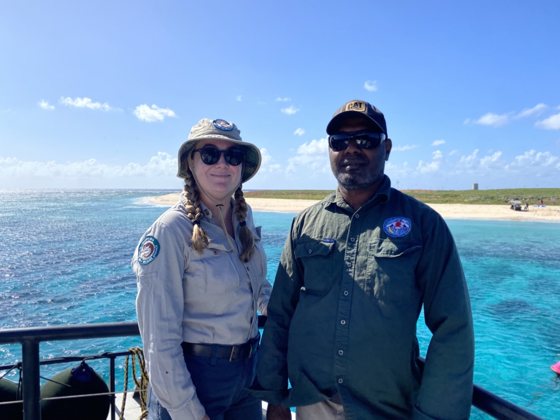 A photo of a Wuthathi Custodian and a Reef Joint Field Management Program ranger on the back of a vessel. Behing them, a bright blue see surrounds Raine Island – which has a small sandy bank surrounding vegetation and a small tower can be seen in the distance. 