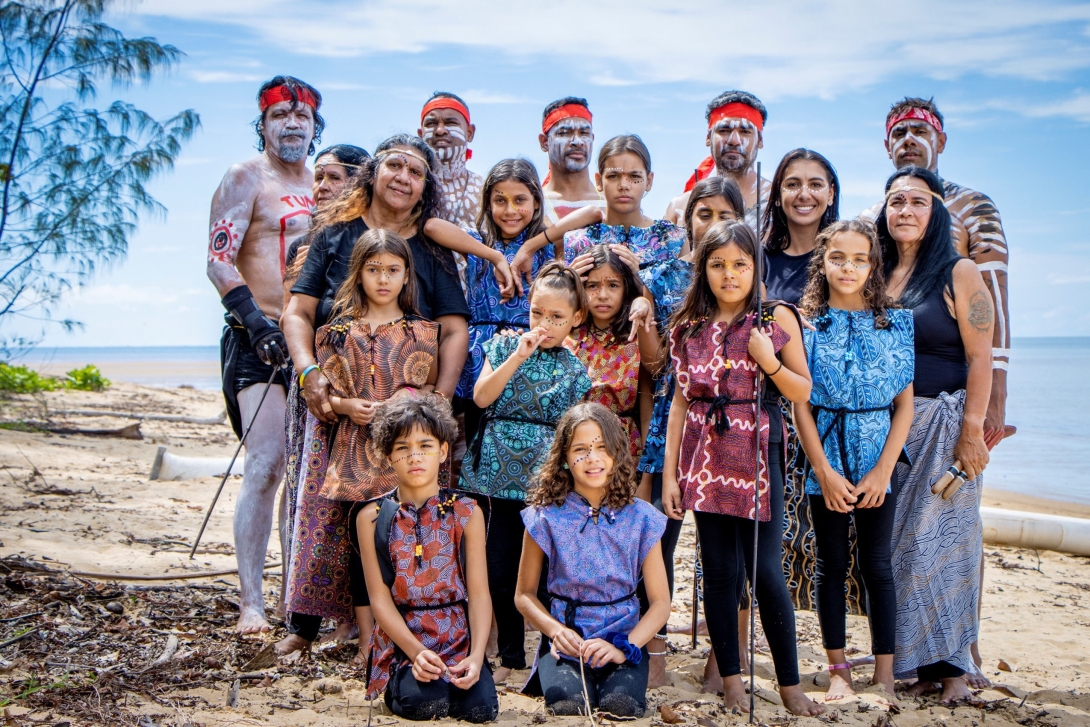 A group of First Nations people standing on a beach. The group is a mix of adults and children. They are dressed for a performance. They look proud and some are smiling. The men in the back row are wearing red headbands and have patterns marked on their bodies with white paint.  One woman is holding a traditional music instrument.