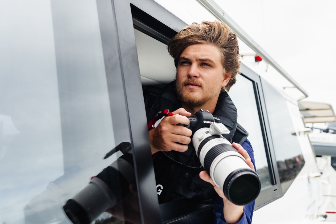 A man looking out the window of a boat holding a camera with a long zoom lens.